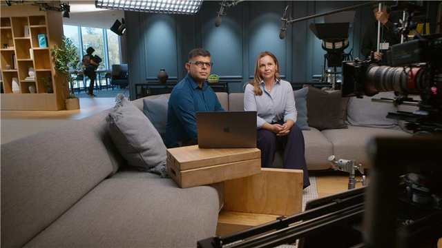 Two people sitting on a couch in an office surrounded by cameras for an interview