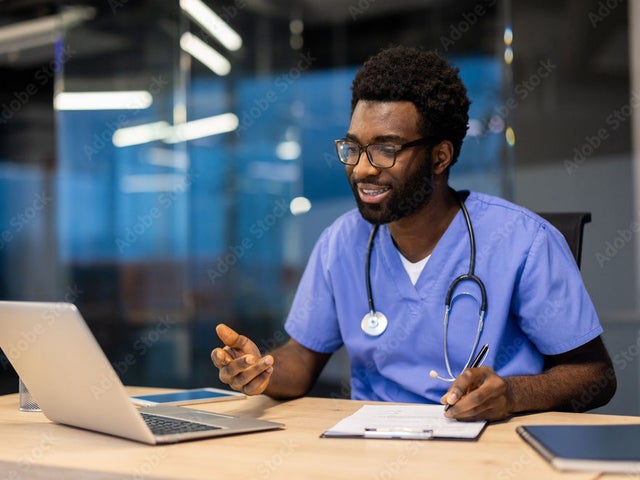Man in scrubs with a stethoscope around his neck working on a laptop at night