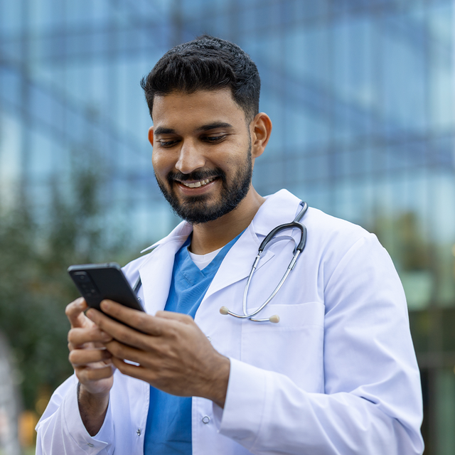 Man wearing scrubs and a lab coat smiling at his phone outdoors in front of a tall building