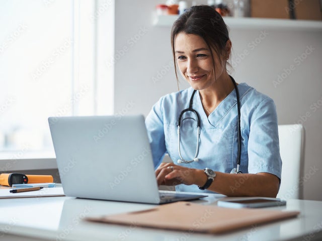 Woman in scrubs with a stethoscope around her neck working on a laptop near a window