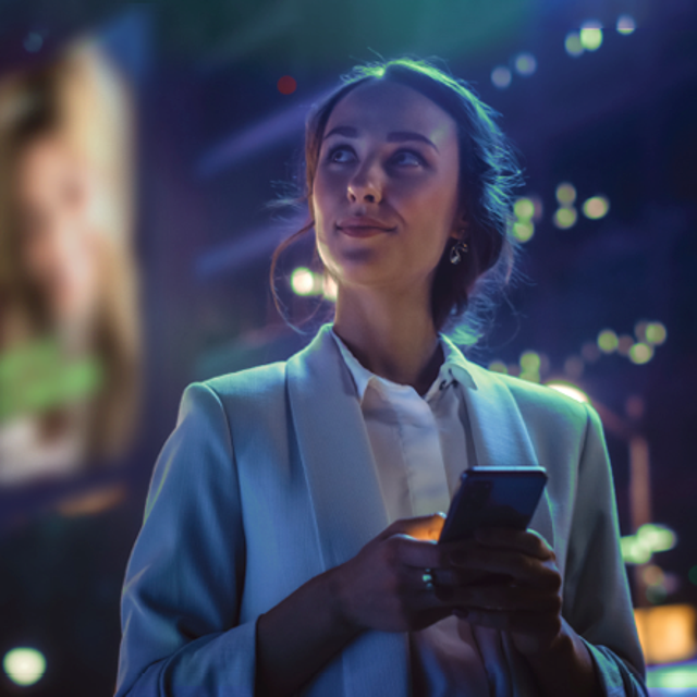 Woman holding a phone outdoors looking up and away in front of tall lit buildings on a dark night