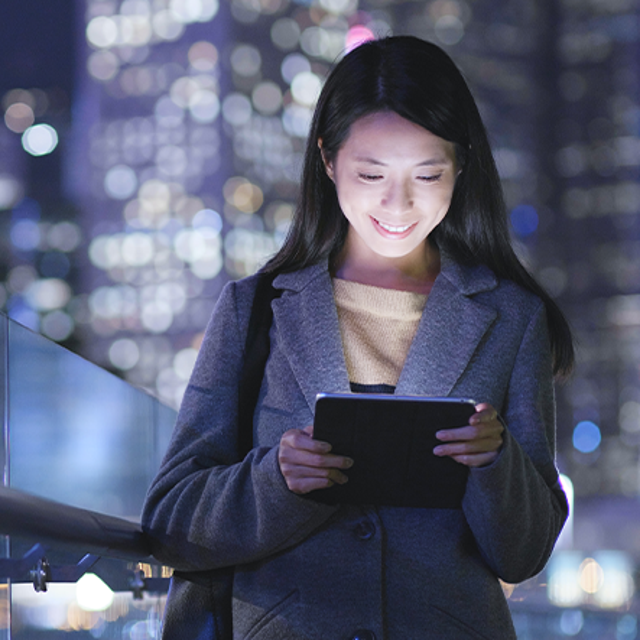 Woman smiling at a tablet in her hands in front of a lit skyscraper at night