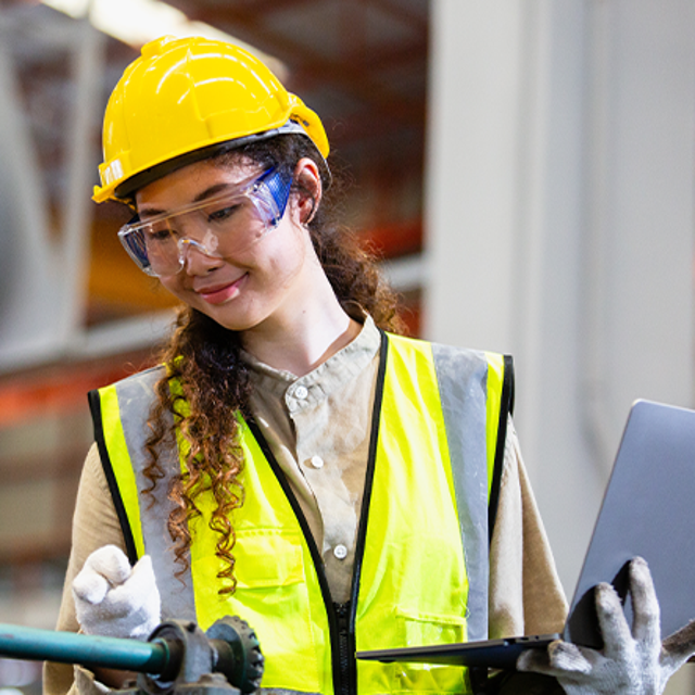 Woman in a hard hat and safety vest holding a laptop near machinery in a factory