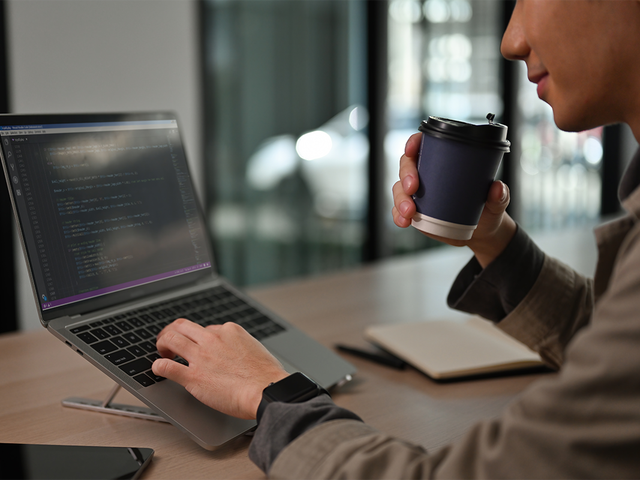 Developer typing on a laptop while holding a cup in one hand
