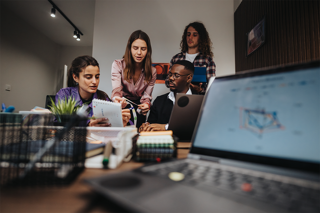 Four workers collaborating behind an open laptop in the foreground