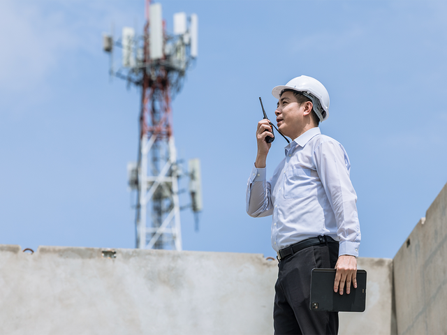Man wearing a hard hat using a walkie-talkie in front of a communications tower