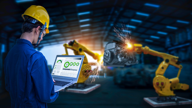 Man wearing a hard hat working on a laptop in front of robotic machines