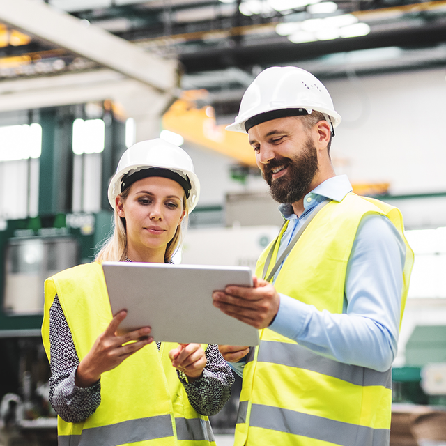 Two workers on a manufacturing floor smiling at a tablet 