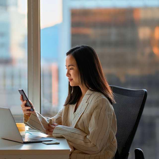 Woman sitting at a desk in front of office window in a high-rise building looking at her phone