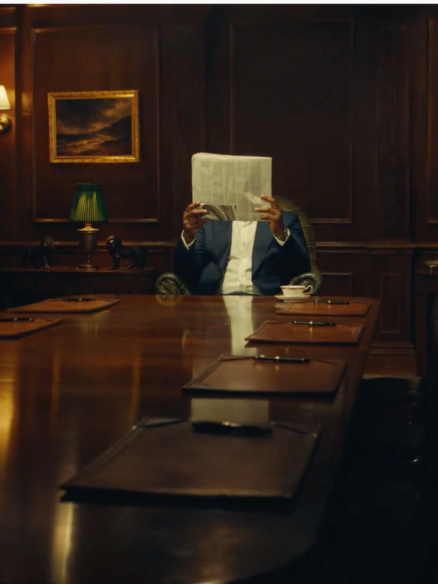 Idris Elba with a newspaper in front of his face at the head of a conference table