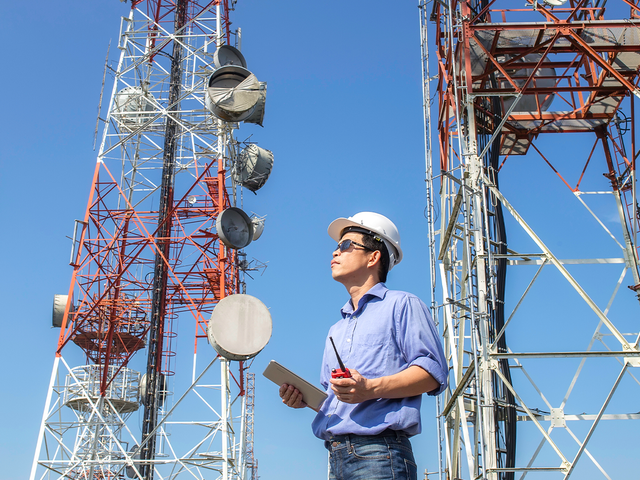 Man wearing a hard hat and holding a tablet surrounded by cell towers