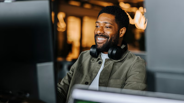 Man with headphones around his neck smiling at his computer monitor