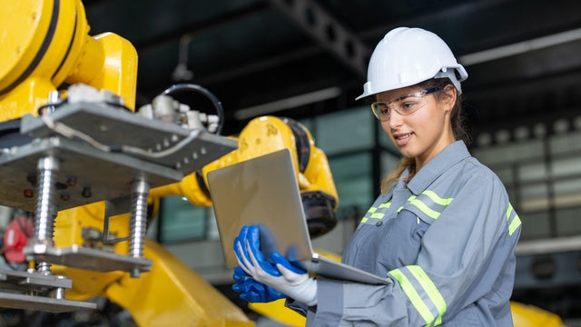 Woman smiling at a laptop while standing next to a machine on a manufacturing floor