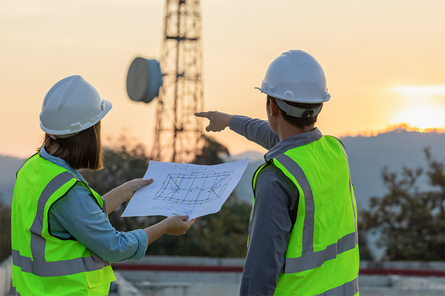Two men in hard hats and construction vests checking a blueprint and pointing to a cell tower in the distance
