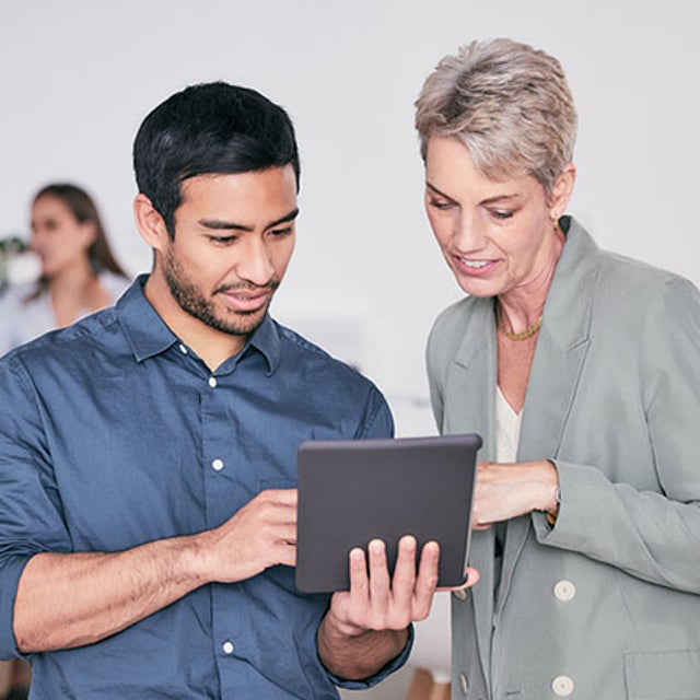 Two workers looking at a tablet while other workers chat at a table