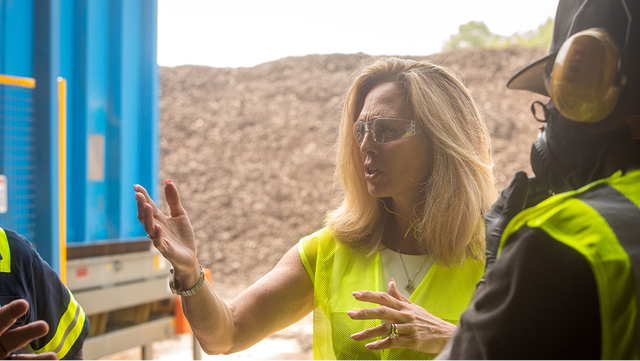 A woman in a safety vest gestures while speaking with a colleague outdoors, with a large pile of recycled tire material visible in the background.