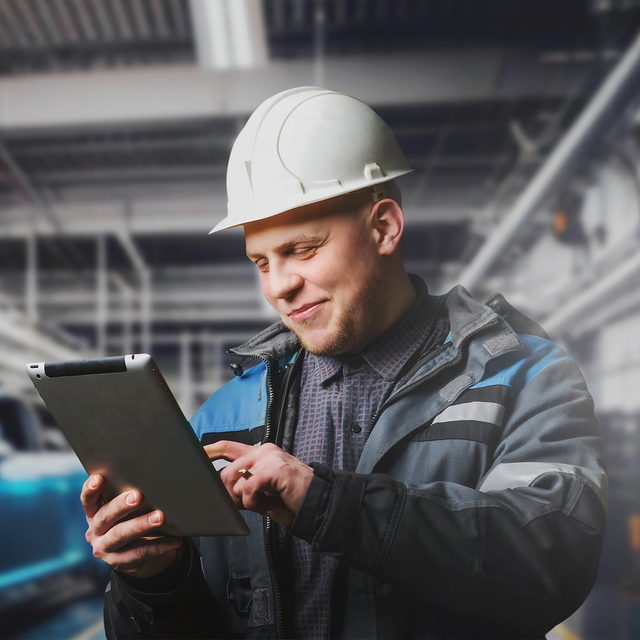 An automotive worker wearing a hard hat and smiling at a tablet in his hands