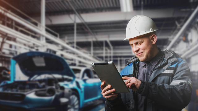 An automotive worker wearing a hard hat and smiling at a tablet in his hands