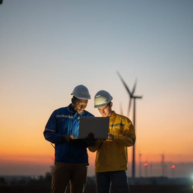 Two field workers looking at a laptop at sunset against a background of windmills