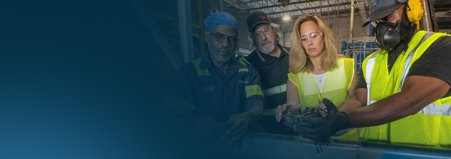 showing four workers in safety vests and gear examining recycled material on a factory floor